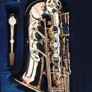 Zlatý saxofon v modrém polstrovaném kufru. Vedle nástroje je umístěn zlatý mouthpiece s korkem a malý plastový sáček s příslušenstvím. Na nástroji ne…