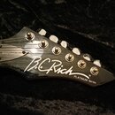 Detail hlavy černé elektrické kytary s nápisy WARLOCK, B.C Rich TM a PLATINUM Series. Na hlavě jsou umístěny šest kovových ladiček. Povrch je lesklý…