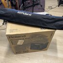 Černý textilní obal na nástroj s bílým logem a nápisem Roland. Obal leží na kartonové krabici, na které je nápis Roland a model KC-150. Na krabici je…