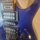 Detailní pohled na modré tělo elektrické kytary s kovovými strunami a hmatníkem z tmavého dřeva. Nástroj má dva humbuckery a jeden jednosnívací sníma…