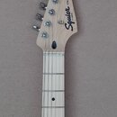 Fotografie zachycuje hmatník a hlavu elektrické kytary z přírodního světlého dřeva. Na hlavě jsou vidět nápisy Squier by Fender a STRATOCASTER®. Povr…