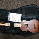 Hnědý dřevěný ukulele v černém polstrovaném obalu. Nástroj je připevněn k obalu černým popruhem, vedle něj leží zapakovaný náhradní set strun. Na hma…