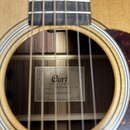 Detailní pohled do otvoru akustické kytary se světlým dřevem desky. Uvnitř je vidět štítek s nápisy Cort GUITAR, MODEL: EARTH200F ATV SG a SERIAL NO:…