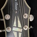 Detail hlavy černé elektrické kytary s bílým lemováním. Na horní části je nápis Blues Lower a v dolní části logo Epiphone. Uprostřed hlavy je vložka…