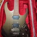 Elektrická kytara s tmavě šedým, kovovým povrchem v červeném tvrdém kufru. Nástroj má dva humbuckery, tremolo s páčkou a čtyři ovládací knoflíky. Pov…
