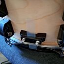 Detailní záběr na spodní část dřevěného bubnu světlé barvy. Na korpusu jsou připevněny černé kovové držáky a kůži drží černý plastový lem. Mezi dvěma…