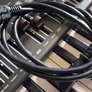 Černý kabel s konektory, který leží smotaný přes klávesy a ovládací panel elektronického nástroje. Na jednom z konektorů je ručně napsaný bílý text V…