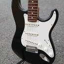 Černá elektrická kytara typu Stratocaster s bílým pickguardem a hnedým hmatníkem. Nástroj má tři bílé snímače, jeden bílý přepínač a tři bílé oválné…