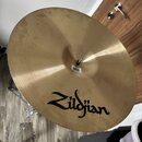 Zlatý kovový činel umístěný na chromovaném stojanu. Na povrchu je černý nápis Zildjian. U středu činelu je viditelný malý bílý papírový štítek s čáro…