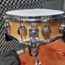Dřevěný snare drum s přírodním povrchem a chromovaným kováním, umístěný na chromovaném stojanu. Na bubnu jsou vidět chromované napínací šrouby a mech…