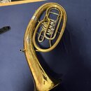 Mosazný železný nástroj typu euphonium v zlaté barvě položený na modré podložce. Na trubici je vidět malý světle šedý štítek s textem, který není pln…