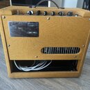 Kytarový kombozesilovač v žlutém texturovaném potahu s hnědým úchytem. Na zadní straně je kovový štítek s nápisy Blues Junior, FENDER MUSICAL INSTRUM…