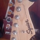Záběr na hlavu elektrokitary z přírodního světlého dřeva. Na povrchu je černý nápis Squier BY FENDER a STRATOCASTER®. Jsou vidět šest chromovaných la…