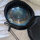 Modrý kovový steel tongue drum umístěný v černém polstrovaném přenosném pouzdře. Na boku nástroje jsou bílé nápisy G, G, F, A, O, D. Vedle pouzdra le…