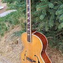 Akustická archtop kytara s hnědým dřevěným povrchem a f-otvory. Na hlavě hmatníku je vidět bílý kosočtvercový ornament. Nástroj stojí na černém stoja…