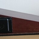Hnědý dřevěný podstavec klavíru nebo syntezizátoru s šikmým horním povrchem. Na boční straně je přišroubovaná černá plastová dvířka s vyraženým kulat…