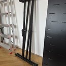 Černý kovový stojan na klávesové nástroje s A-tvarem a teleskopickými nohami. Stojan je umístěn v interiéru na dřevěné podlaze vedle hliníkového žebř…