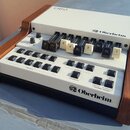 Bílý elektronický klávesový syntetizátor s dřevěnými bočními panely. Na horním panelu je nápis OB-3 a logo Oberheim, v dolní části je opět logo Oberh…