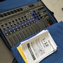 Šedý digitální mixážní stolek Zoom L-12 LiveTrak umístěný v modrém transportním pouzdře. Zařízení má řadu ovládacích prvků, posuvníků a nápisy jako M…