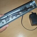 Fotografie zachycuje zadní stranu šedého elektronického zařízení s připojeným kabelem a samostatný černý síťový adaptér. Na šasi je vidět nápis TRUE…