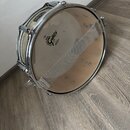 Stříbrný snare drum značky Gretsch s béžovými membránami. Na spodní membráně je černé logo Gretsch a nápis REMO. Nástroj má chromované obruče a mecha…