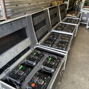 V otevřených transportních kufrech z hliníku a černého překlijení je umístěno několik černých rectángulových zařízení s madly. Na zadních panelech př…