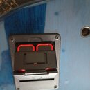 Detailní pohled na černý plastový konektor pro kabel, který je přišroubován k modrému povrchu nástroje. Konektor má červené vnitřní prvky a na horní…