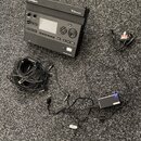 Černý elektronický modul s nápisy Roland a V-Drums, vybavený displejem, ovládacími tlačítky a knoflíkem. K modulu patří černý napájecí adaptér s nápi…
