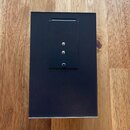 Černý obdélníkový box z matního materiálu položený na dřevěném povrchu. V horní části je umístěna menší černá destička s dvěma zlatými kovovými kolík…
