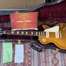 Zlatá elektrická kytara typu Les Paul v černém tvrdém kufru s červeným plyšovým vnitřkem. Na hlavě kytary je bílý nápis Gibson a podpis. V kufru se n…