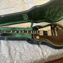 Tmavně hnědá elektrická kytara typu Les Paul s krémovým pickguardem a dvěma humbuckery v černém tvrdém kufru s zeleným polstrováním. Na hmatníku jsou…