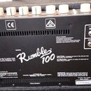 Zadní strana černého kytarového komba s bílými oválnými knoflíky. Na černé kovové desce je bílý nápis Rumble 100, značka CE a symboly varování. Jsou…