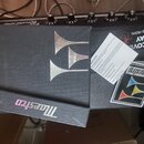 Detailní pohled na černý efekt pedál s texturou kůže. Na horní části je stříbrné logo ve tvaru tří rohů a nápisy DISCOVER DELAY EFFECTS PEDAL. V doln…