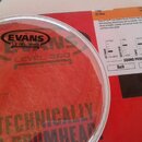 Průhledná plastová bubna v bílém obruči položená na červeném kartonovém obalu. Na bubně je černý nápis EVANS G2 LEVEL 360 A D'ADDARIO INNOVATION. Na…