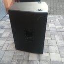 Dva černé, hranaté reproduktorové boxy umístěné na dlažbě. Přední box má na horní hraně dvě kovové úchytky a na boku panel s konektory. Povrch je mat…