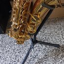 Zlatý saxofon umístěný na černém kovovém stojanu. Na čáře nástroje je viditelný neúplný nápis, ze kterého jsou čitelné znaky RR A a S 6. Povrch nástr…
