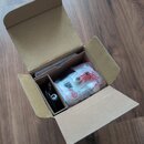Otevřený hnědý kartonový krabici s vnitřními přihrádkami. Uvnitř je umístěn černý předmět a plastový sáček s bílým obalem, na kterém je červený nápis…