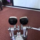 Fotografie zachycuje detail dvojpedálového mechanismu bicích pedálů v kovovém provedení. Pedály mají černé plastové špičky s bílými plstěnými nebo lá…