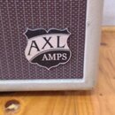 Detailní záběr na roh kytarového komba s hnědou textilní maskou a krémovým rámem. Na masce je umístěno logo ve tvaru štítu s nápisem AXL AMPS. Povrch…