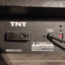 Detail zadní strany černého zesilovače s bílým nápisem TNT. Na přístroji je vidět napájecí kabel, vypínač a štítek s textem 220-230/240V~ 50/60 Hz 50…