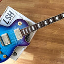 Modro-fialová elektrická kytara s typem korpusu Les Paul a plameníjím dřeva. Na hlavě hmatníku je nápis "Gibson" a logo "Les Paul". Kytara má dva hum…