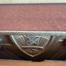Detailní pohled na zlatou kovovoudesku s logem vkládanou do nástroje. Na desce je vyražen nápis PETROF a nad ním písmena A a P v rámci stylizovaného…