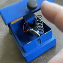 Modrý efekt pedál s otevřeným horním krytem, uvnitř kterého je vidět mechanismus s pružinou a zapojenými červeným a černým kablem. Na vnitřní straně…