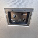 Detail zadní strany reproduktoru s černým ovládacím panelem v šedém rámu. Panel obsahuje regulační knoflík s nápisem HIGH RANGE, dva konektory s popi…