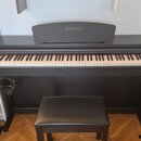 Černý digitální piano v černém dřevěném korpusu s přívodní stoličkou v odpovídající barvě. Na horní části krytu i v pravém dolním rohu je bílý nápis…