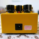 Zadní strana žlutého efektpedálu s třemi černými oválnými knoflíky a malou červenou kontrolkou. U napájecího konektoru je bílá nálepka s nápisem USE…