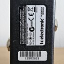 Zadní strana černého efektpedálu z kovu s texturovaným povrchem. Na bílém štítku je uveden nápis tc electronic®, DESIGNED IN DENMARK, E600002111 a na…
