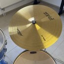 Zlatý činel typu ride s nápisy „zuriet“ a „20" RIDE“. Na levé straně je umístěno logo „Zuriet“. Činel je umístěn nad bílým bubnem, na jehož horním ok…