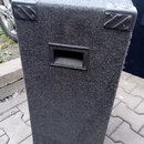 Černý reproduktorový box potažený hrubým textilem. V horní části jsou umístěny dvě kovové rohové krytky a uprostřed je plastová rukojeť. Povrch texti…