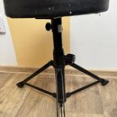 Černý pianistický taburet s kulatým polstrováním z umělé kůže a tříramenou kovovou základnou. Na horní části nohy je viditelný bílý štítek s textem,…
